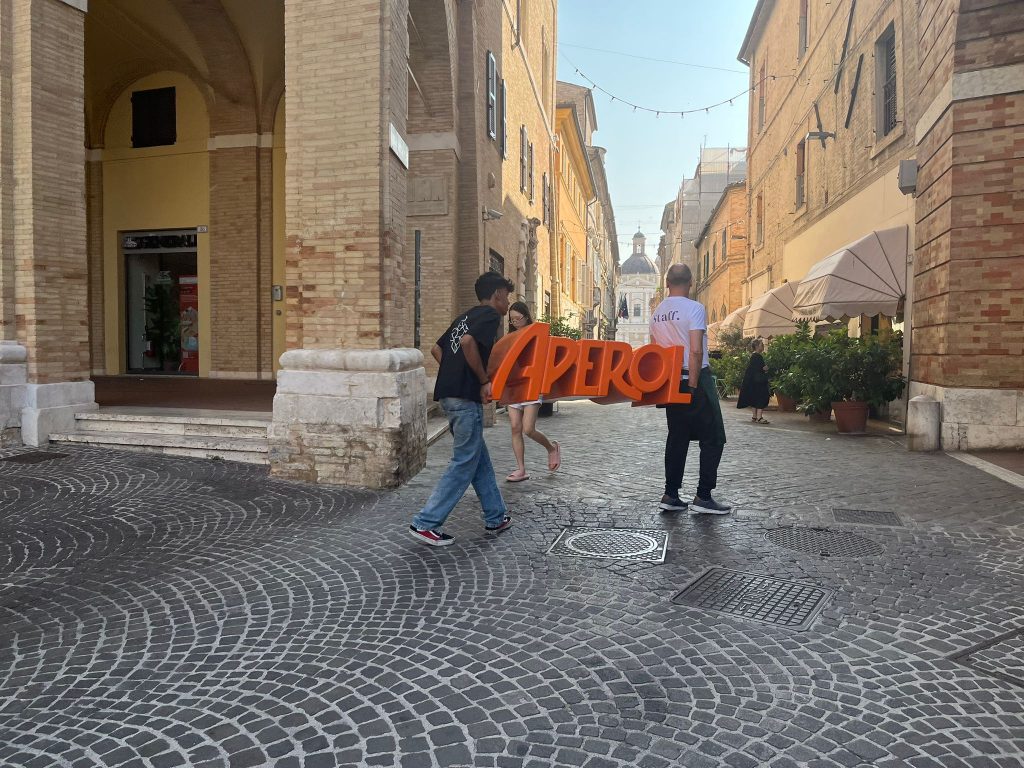 Image où deux hommes portent un insigne Aperol, marque italienne indispensable à la préparation des Spritz. Ils se trouvent dans la rue parallèle à la place principale, où deux nombreux établissement proposent des spritz et pizza à Macerata.